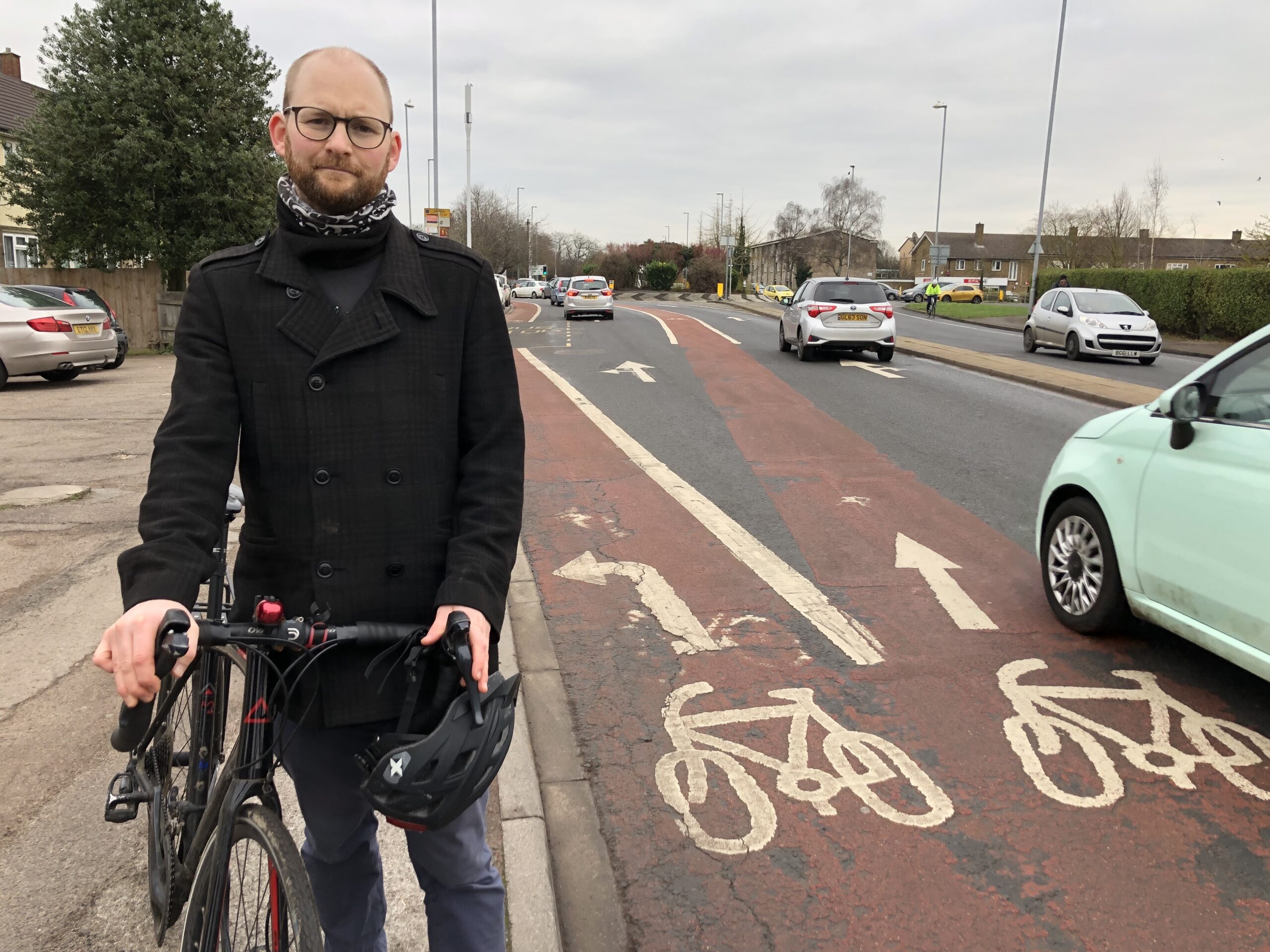 Councillor Matt Howard stands with his bicycle by a busy road with multiple lanes for cars and cycles