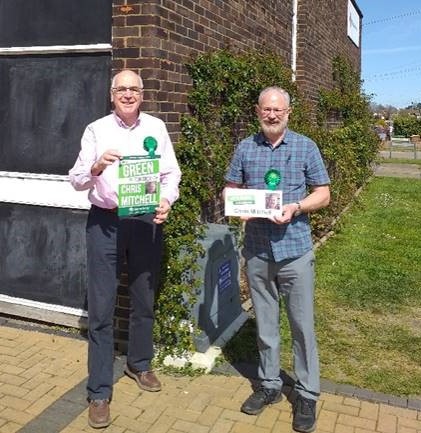 Andrew and Chris stand outside the Red Cross Centre in the sun. They are wearing Green Party rosettes and are holding leaflets.