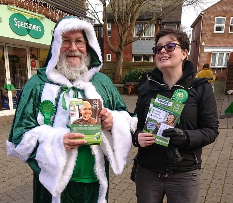 Bobbie Curran stands with a person dressed as Green Santa on Tring High Street. They both wear Green Party rosettes and hold flyers.